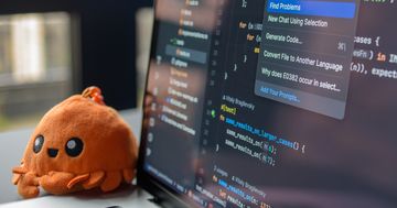 A laptop screen showing a code editor with a cute orange crab plush toy beside it.