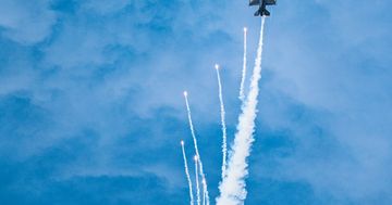 Military jet releasing flares during an impressive aerial maneuver against a bright blue sky.